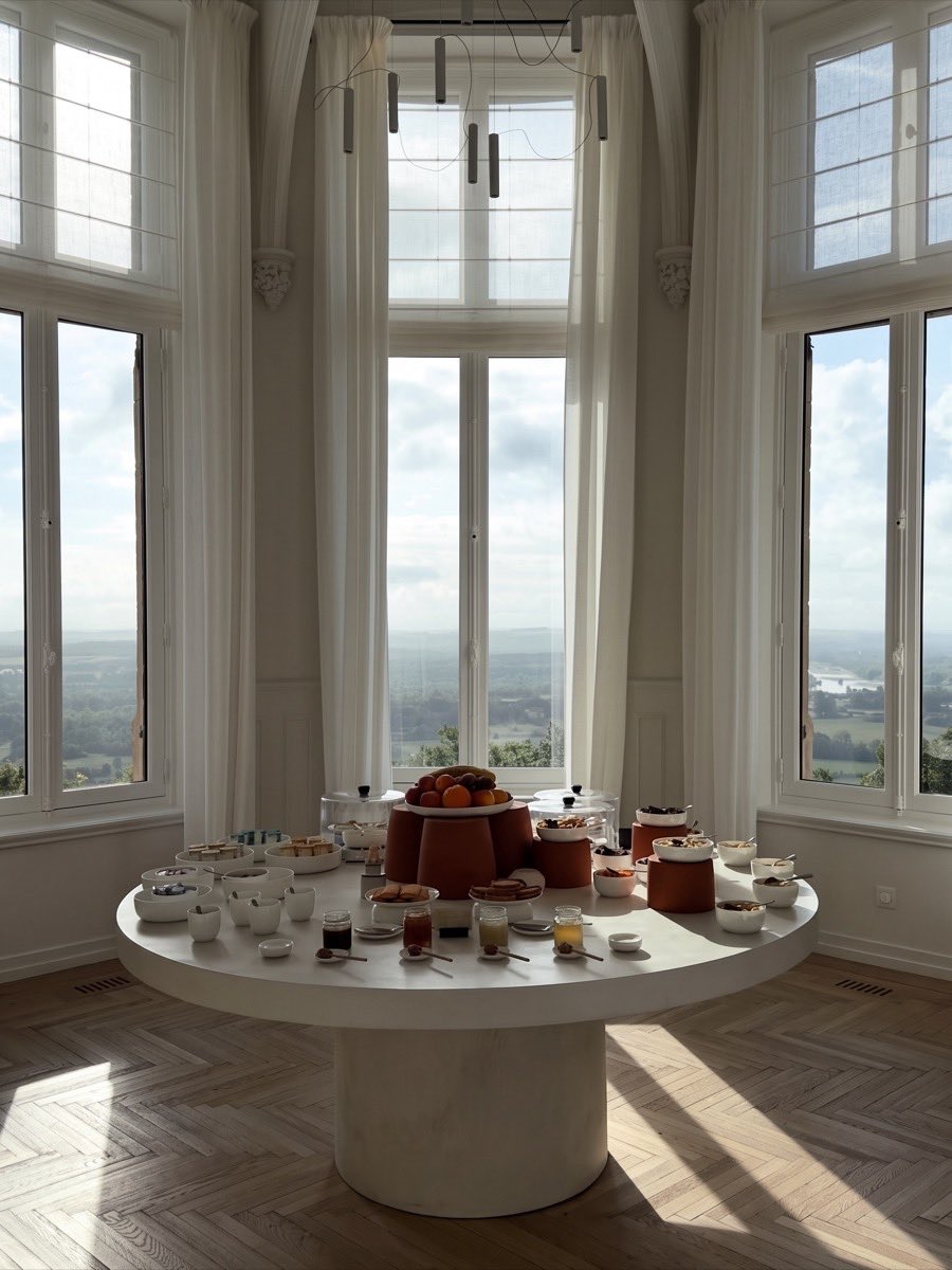 Le petit-déjeuner d’un hôtel : une table ronde dressée dans une baie de hautes fenêtres ouvertes sur une vallée.
