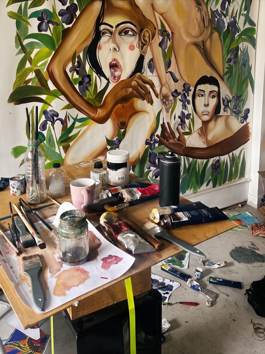 L’atelier d’une peintre : une grande toile en cours au mur, une table couverte de pinceaux, de tubes et de pots.